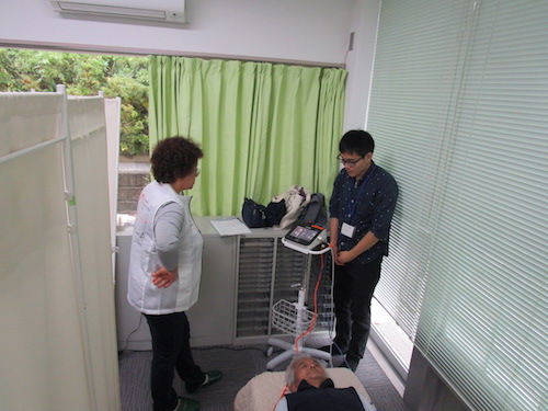 血圧測定中の参加者の近くで差し向かいで立つ看護師のミキちゃんと学生ボランティア